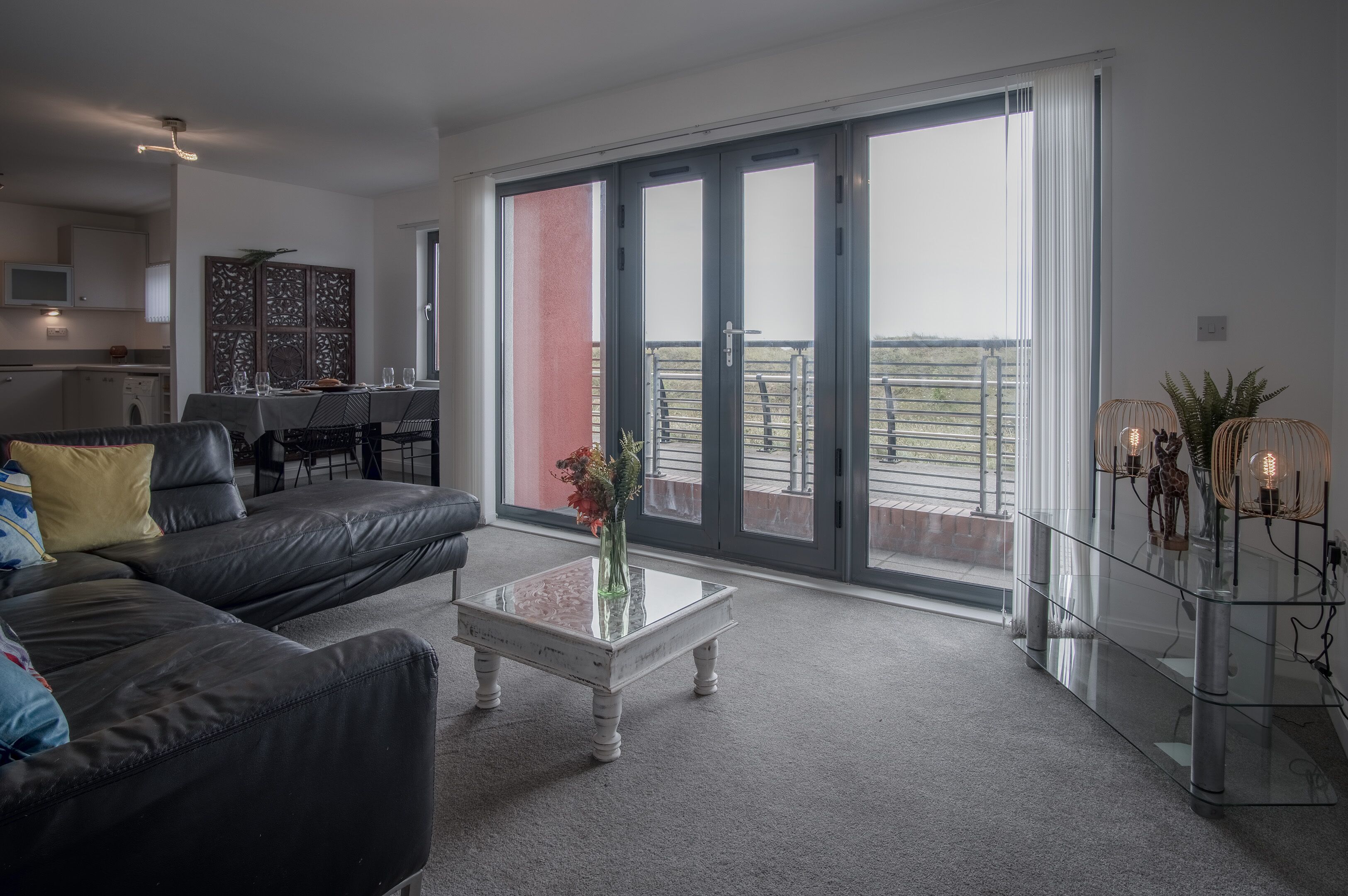 A modern living room with a black leather sofa, a glass coffee table, and a dining area with a table set for two.