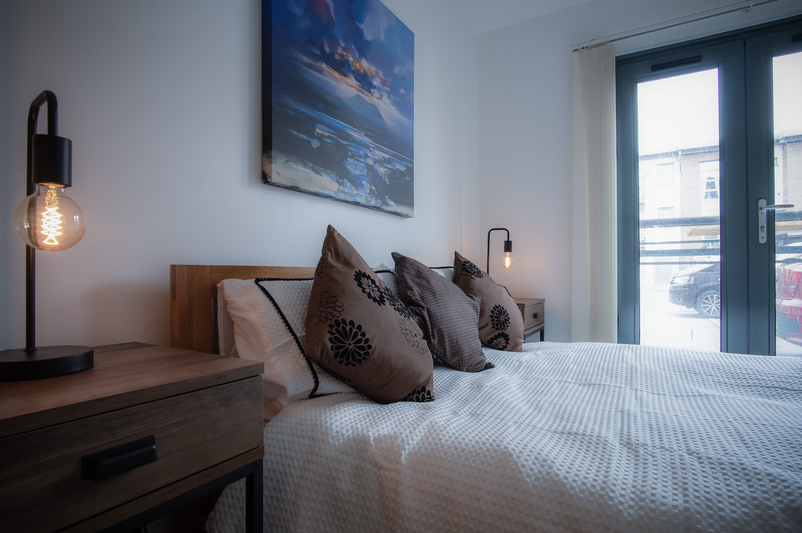 A bedroom with a bed, pillows, a nightstand, a lamp, and a painting on the wall.