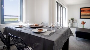 Un coin repas avec une table dressée pour deux personnes, une vue sur la mer à travers de grandes fenêtres et un tableau accroché au mur.