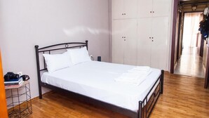 A single-bed room with a wooden floor, a metal bed frame, white bedding, a small bedside table with a camera, and a wardrobe with multiple doors. - Fabulous Apartment in Pireaus (Piraeus)