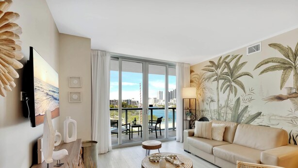 A modern living room with a sofa, a television, and a balcony with a city view. - Modern Resort : Balcony+ Gym+ Amazing Pool (Hallandale Beach)