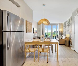 Une cuisine moderne avec un réfrigérateur en acier inoxydable, une table à manger avec des chaises, un luminaire suspendu et une vue sur l’extérieur grâce à de grandes fenêtres.