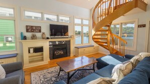 A living room with a fireplace, a television, a blue sofa, a wooden staircase, and a view of a lake.