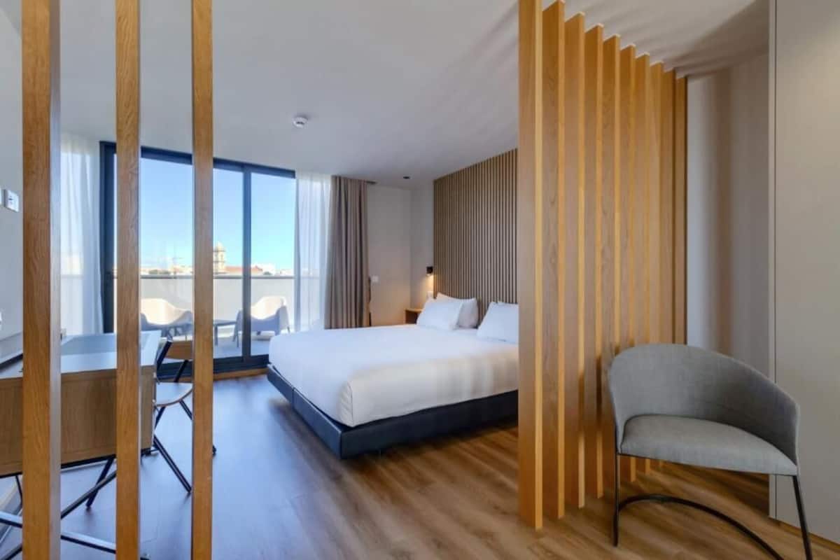 A modern hotel room with a large bed, a desk, and a chair. There is a view of the city through large windows.