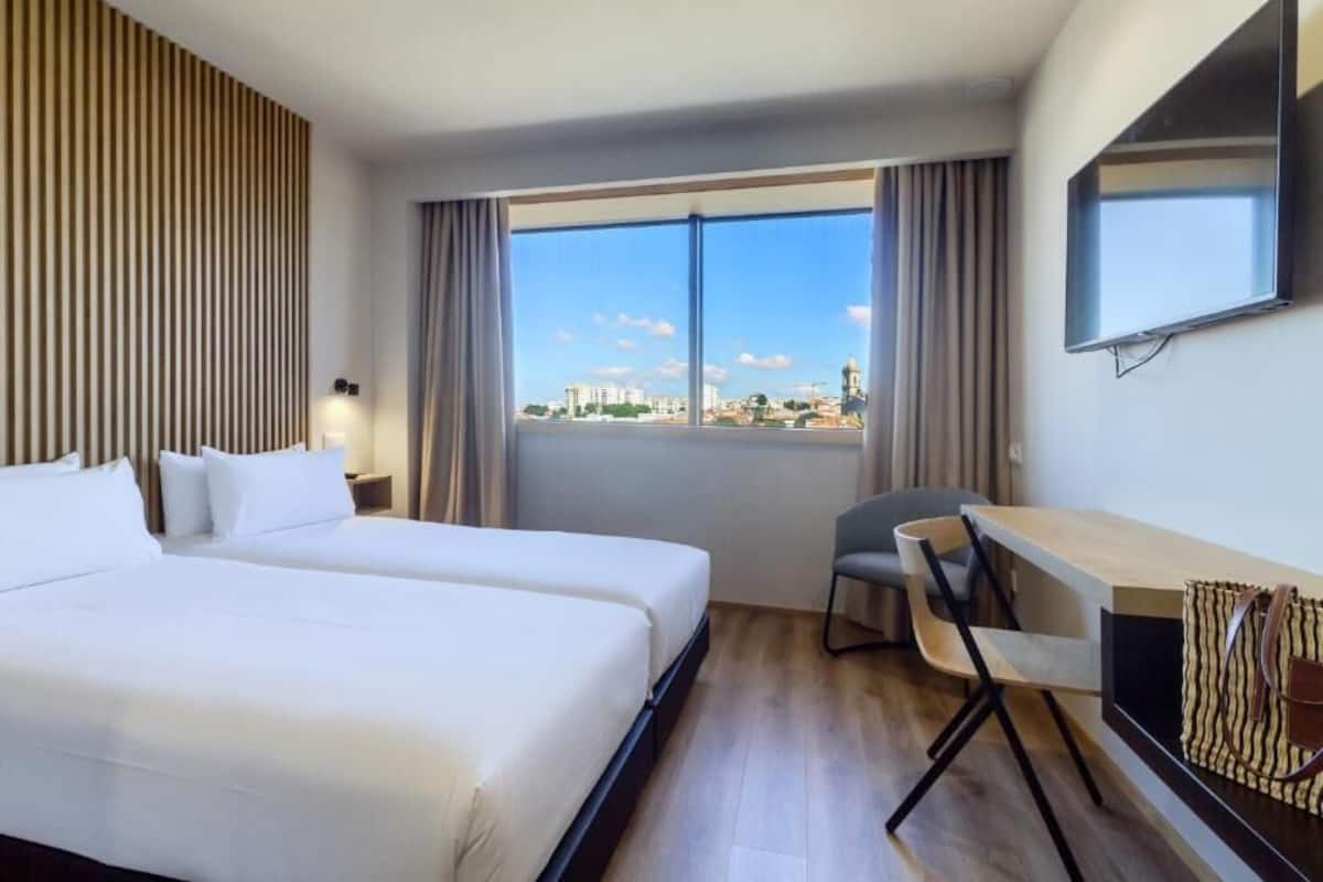 A hotel room with two beds, a desk, a chair, a TV, and a large window with a city view.