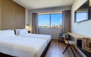 A hotel room with two beds, a desk, a chair, a TV, and a large window with a city view.