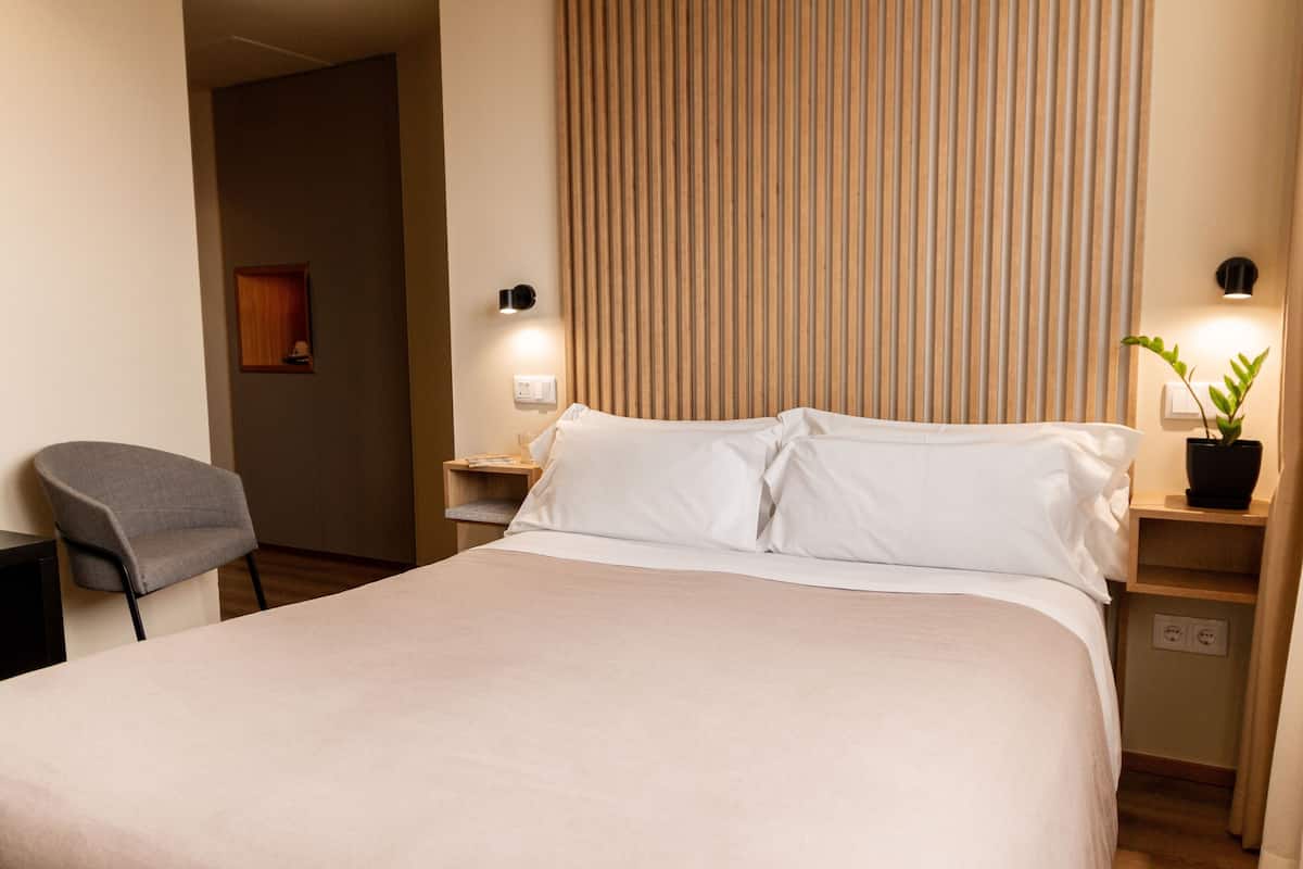 A hotel room with a bed, a chair, a nightstand, a potted plant, and a wall-mounted light fixture.