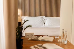 Una camera d'hotel con un letto, un comodino con un libro, una pianta e una bottiglia d'acqua.