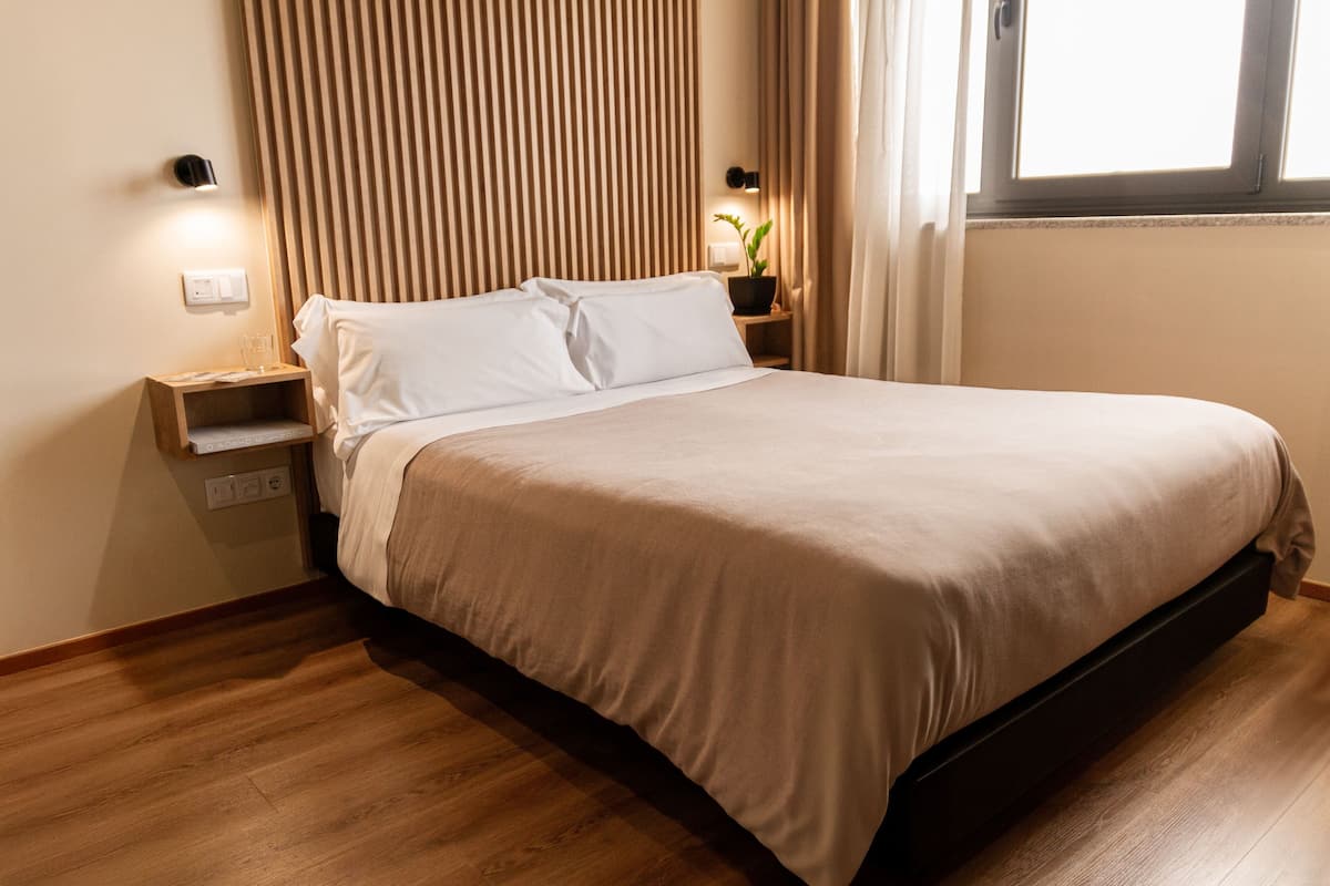 A neatly made bed with a wooden headboard, a bedside table with a lamp, and a window with curtains.