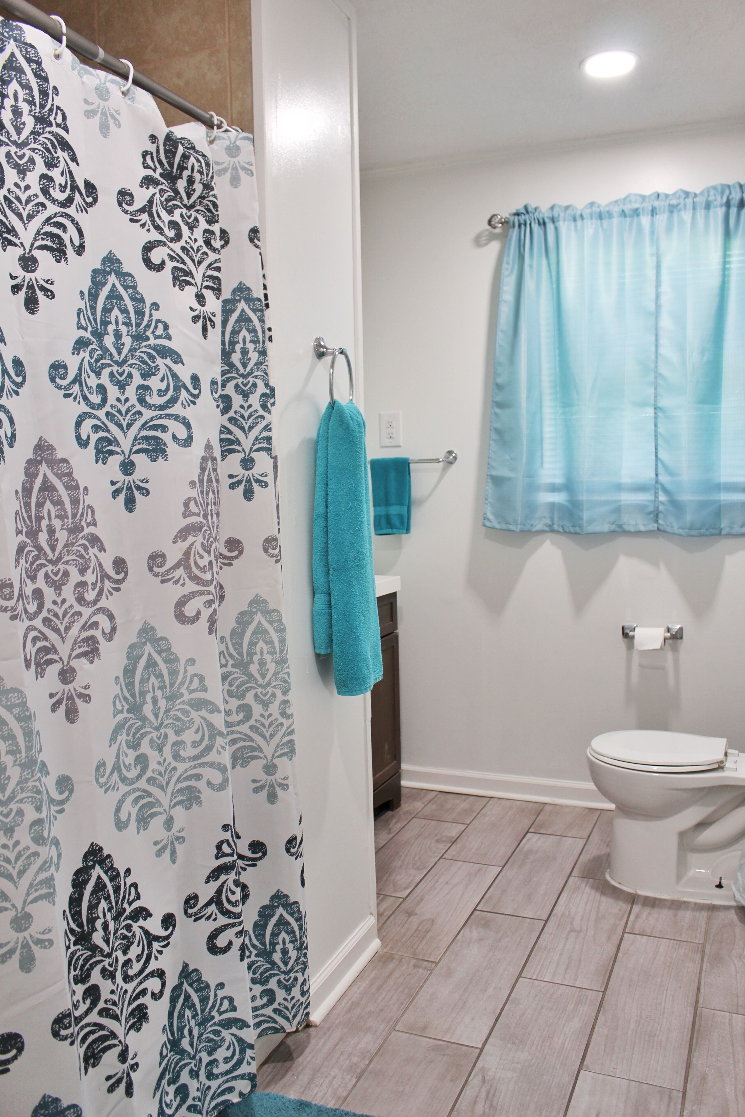 A bathroom with a shower curtain, a towel rack with a towel, a toilet, and a window with a curtain.