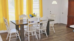 Een eetgedeelte met een houten tafel en witte stoelen, gele gordijnen en een raam met zonwering.