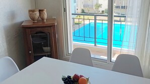 Een eetgedeelte met een tafel gedekt voor twee personen, een fruitschaal en uitzicht op een zwembad door een raam.