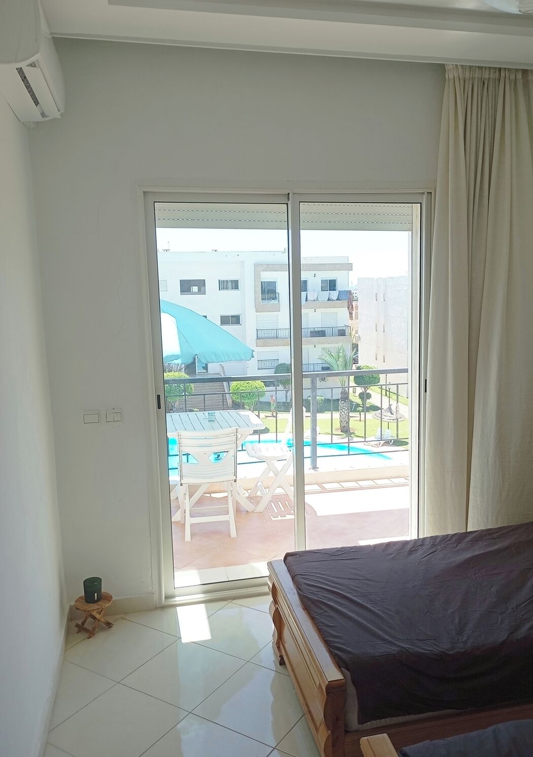 Un dormitorio con cama, una puerta corrediza de vidrio que da a un balcón con piscina y mobiliario de exterior.