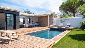 Un área de piscina moderna al aire libre con cubierta de madera, sillones de descanso y un pequeño patio con mesa y sillas.