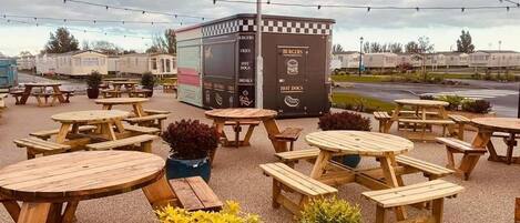 An outdoor seating area with wooden tables and benches, a food truck labeled 'Burger's', and string lights.