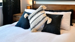 Een bed met wit beddengoed, een gestreept dekentje en twee zwarte kussens met kwastjes.