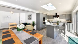 Een moderne keuken met een centraal eiland, een eetgedeelte met een houten tafel en zwarte placemats, en een zithoek met een bank en een plant.