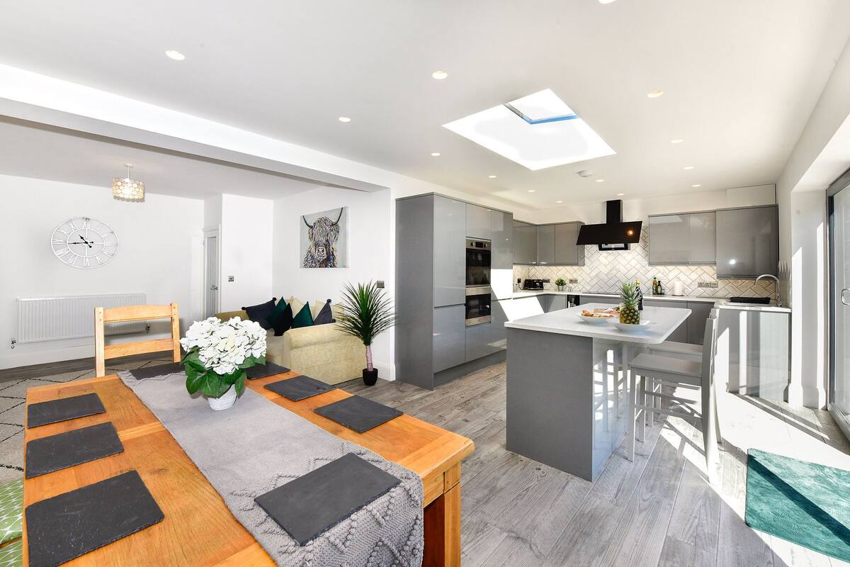 Une cuisine moderne avec un îlot central, un coin repas avec une table en bois et des sets de table noirs, et un espace de vie comprenant un canapé et une plante.