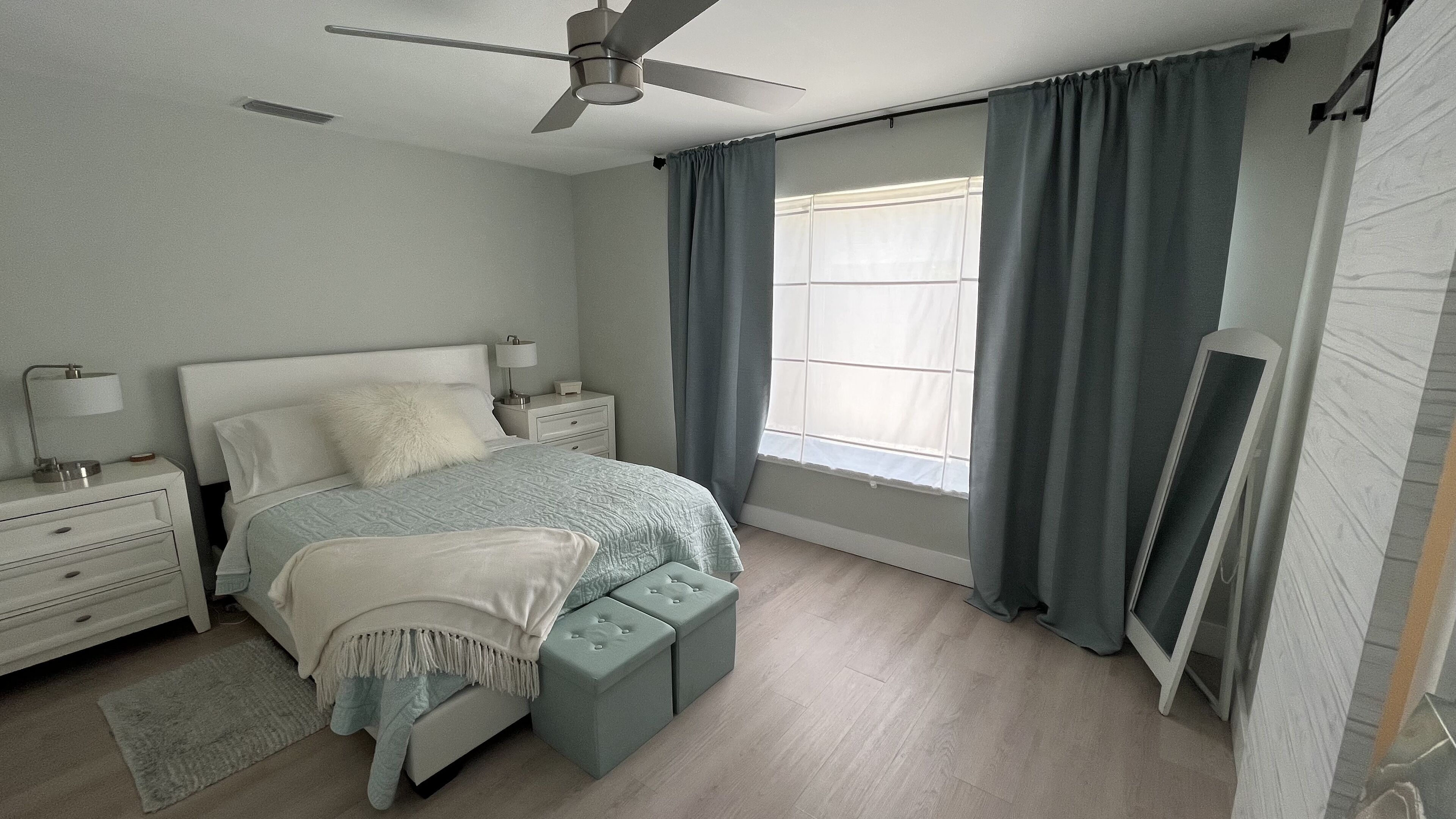 Une chambre à coucher comprenant un lit, des tables de chevet, un ventilateur de plafond, une fenêtre avec des rideaux et un téléviseur fixé au mur.