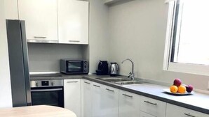 A modern kitchen with white cabinets, a stainless steel refrigerator, a microwave, a kettle, and a sink. There is a dining table with a black chair and a fruit bowl on the counter. - Charming Apartment in Faliro (Palaio Faliro)