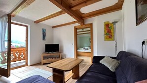A living room with a wooden ceiling, a TV on a wooden stand, a sofa, and a bed in the adjacent room. - Vacation apartment on the farm (Wald)