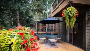 Una terraza de madera con una mesa y sillas, rodeada de exuberante follaje y macetas colgantes de flores.