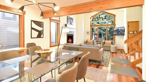 Una sala de estar moderna con una mesa de comedor de vidrio, asientos cómodos, chimenea y ventanas amplias que ofrecen luz natural y vistas.