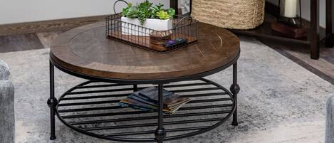 Una mesa redonda de madera con base metálica, una cesta tejida y una vela al fondo.