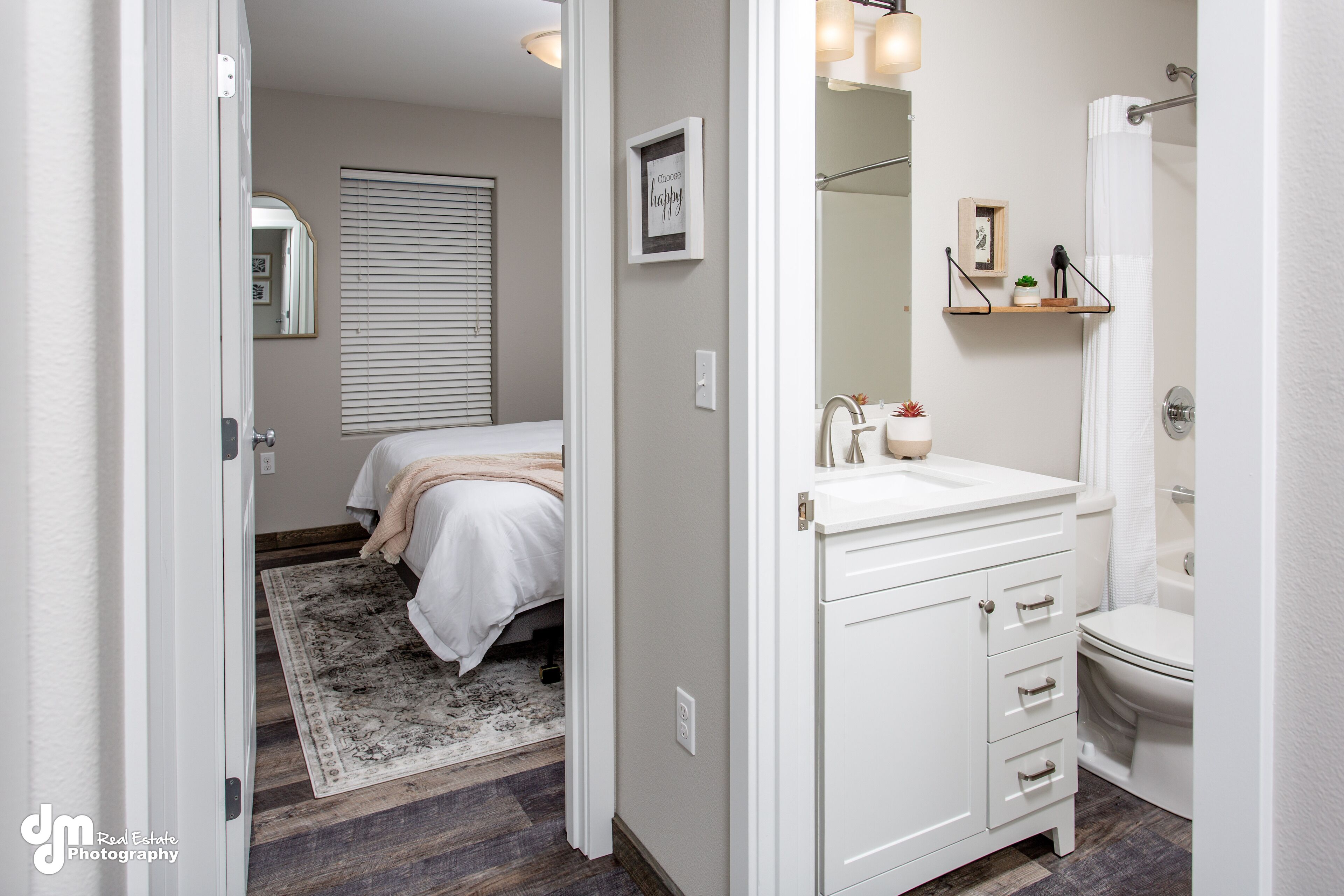 Een slaapkamer met een bed, nachtkastje en een nachttafel. Er is een badkamer met een wastafel, spiegel en toilet. De kamer heeft een houten vloer en een vloerkleed.