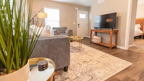 A living room with a grey sofa, a coffee table, a TV on a wooden stand, a potted plant, and a door leading to another room. - Susitna Suites (Wasilla)