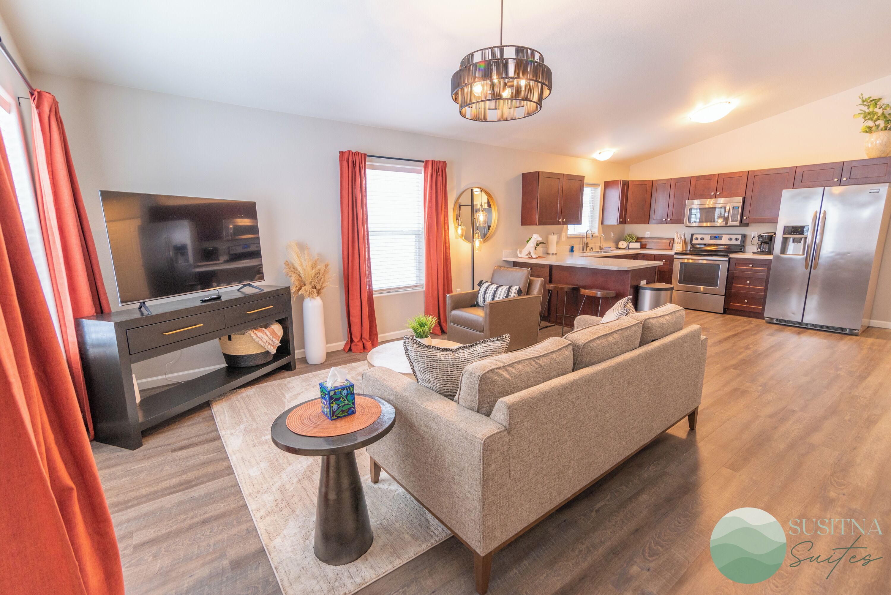 A modern living room with a sofa, TV, and a small table. There is a kitchen area with a refrigerator and microwave.