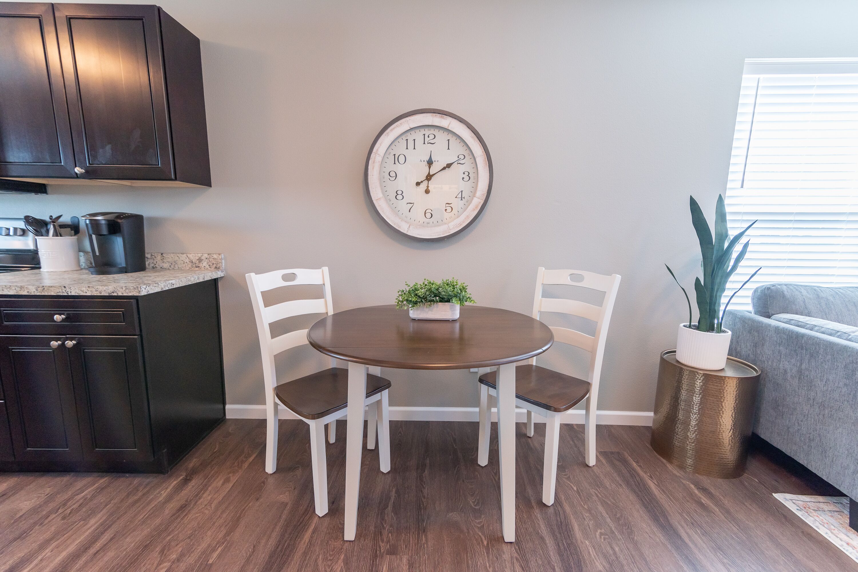 Uma área de refeições com uma mesa redonda, duas cadeiras brancas, um relógio na parede, uma cafeteira e uma planta em uma mesinha lateral.
