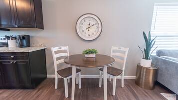 Uma área de refeições com uma mesa redonda, duas cadeiras brancas, um relógio na parede, uma cafeteira e uma planta em uma mesinha lateral.
