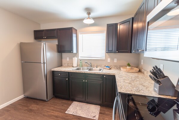 A modern kitchen with dark cabinets, a stainless steel refrigerator, a granite countertop, a built-in oven, and a window with blinds. - Susitna Suites (Wasilla)