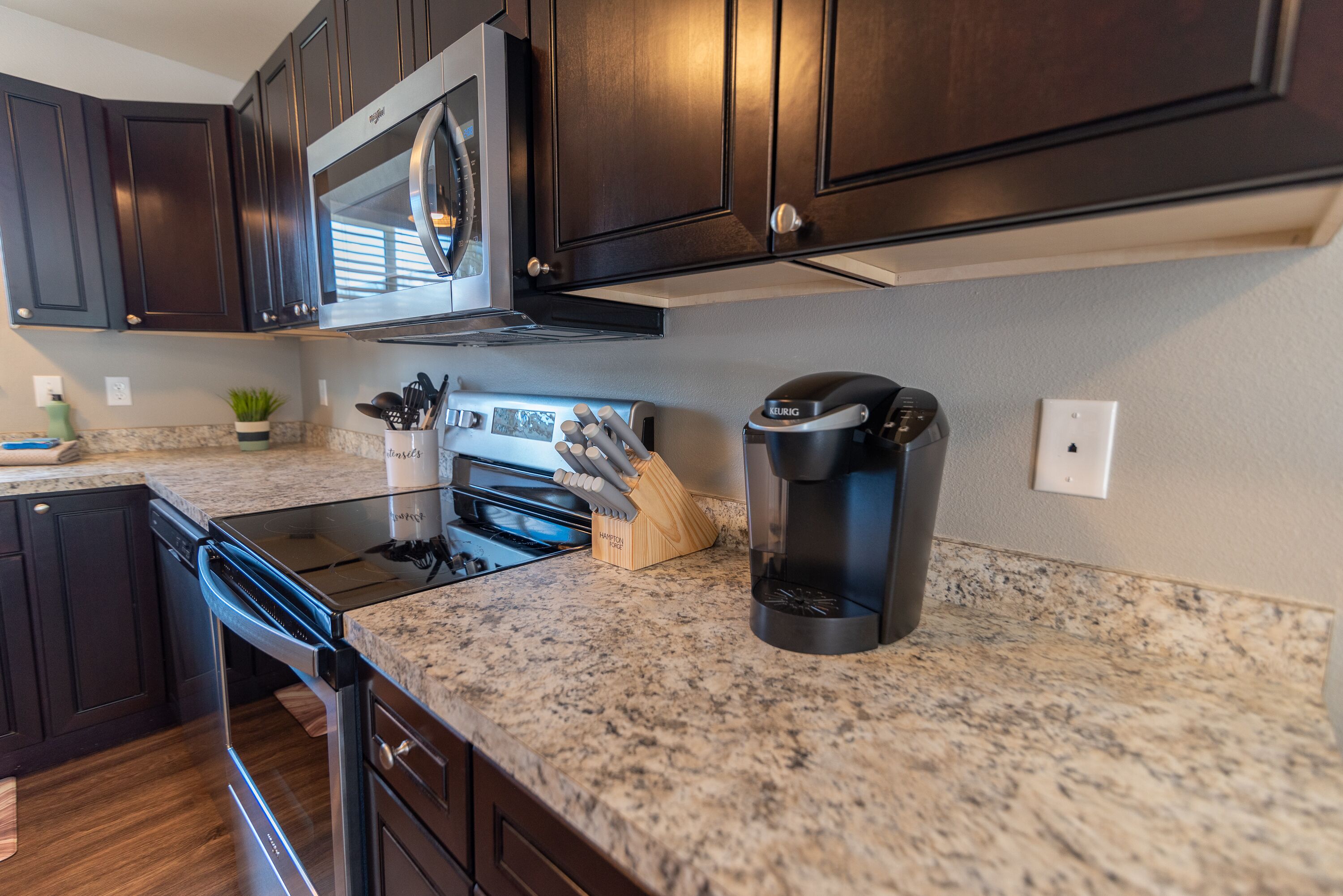 A kitchen with dark wooden cabinets, a stainless steel range hood, a coffee maker, and a knife block on the granite countertop.