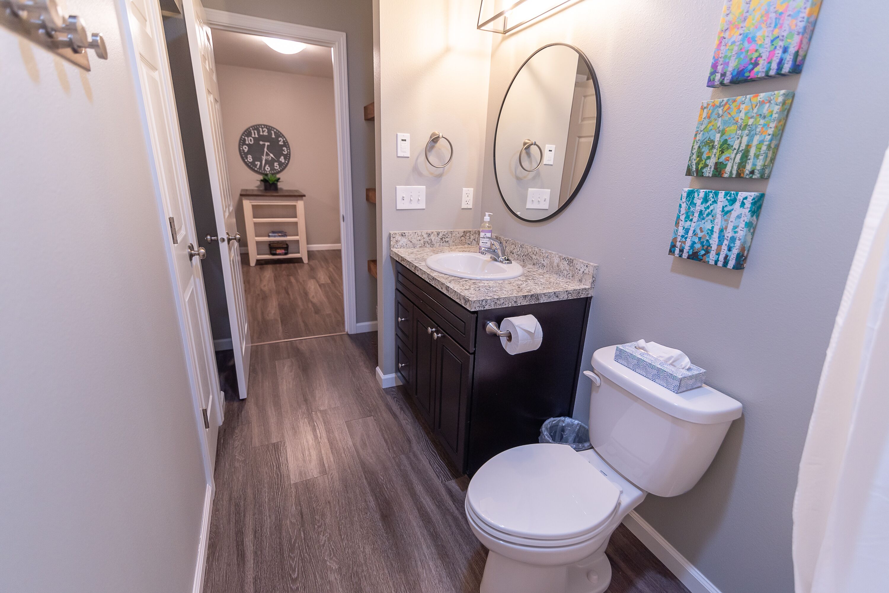 A bathroom with a white toilet, a sink with a marble countertop, a dark vanity, a large mirror, and a clock on the wall.