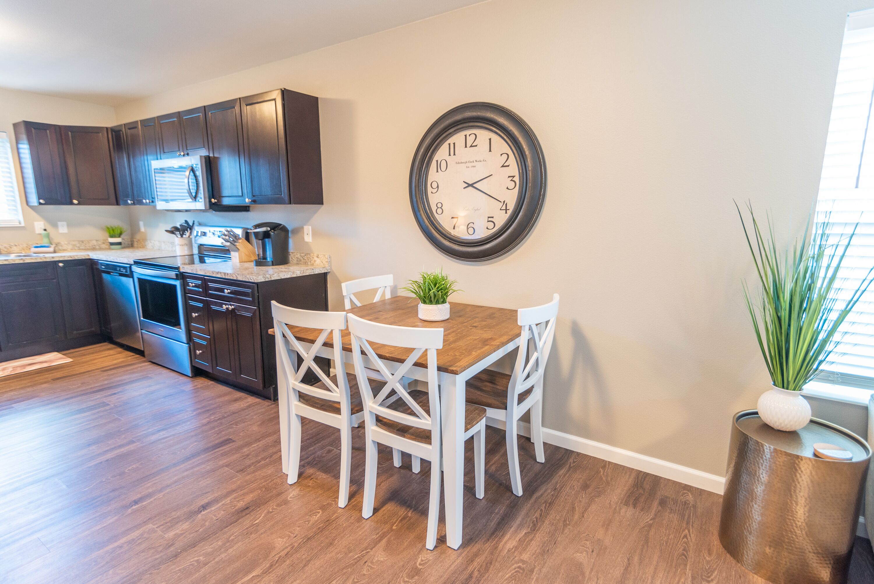 A modern kitchen with dark wooden cabinets, a stainless steel refrigerator, and a wooden dining table with white chairs. A round wall clock is mounted above the table.