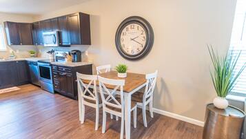 A modern kitchen with dark wooden cabinets, a stainless steel refrigerator, and a wooden dining table with white chairs. A round wall clock is mounted above the table.