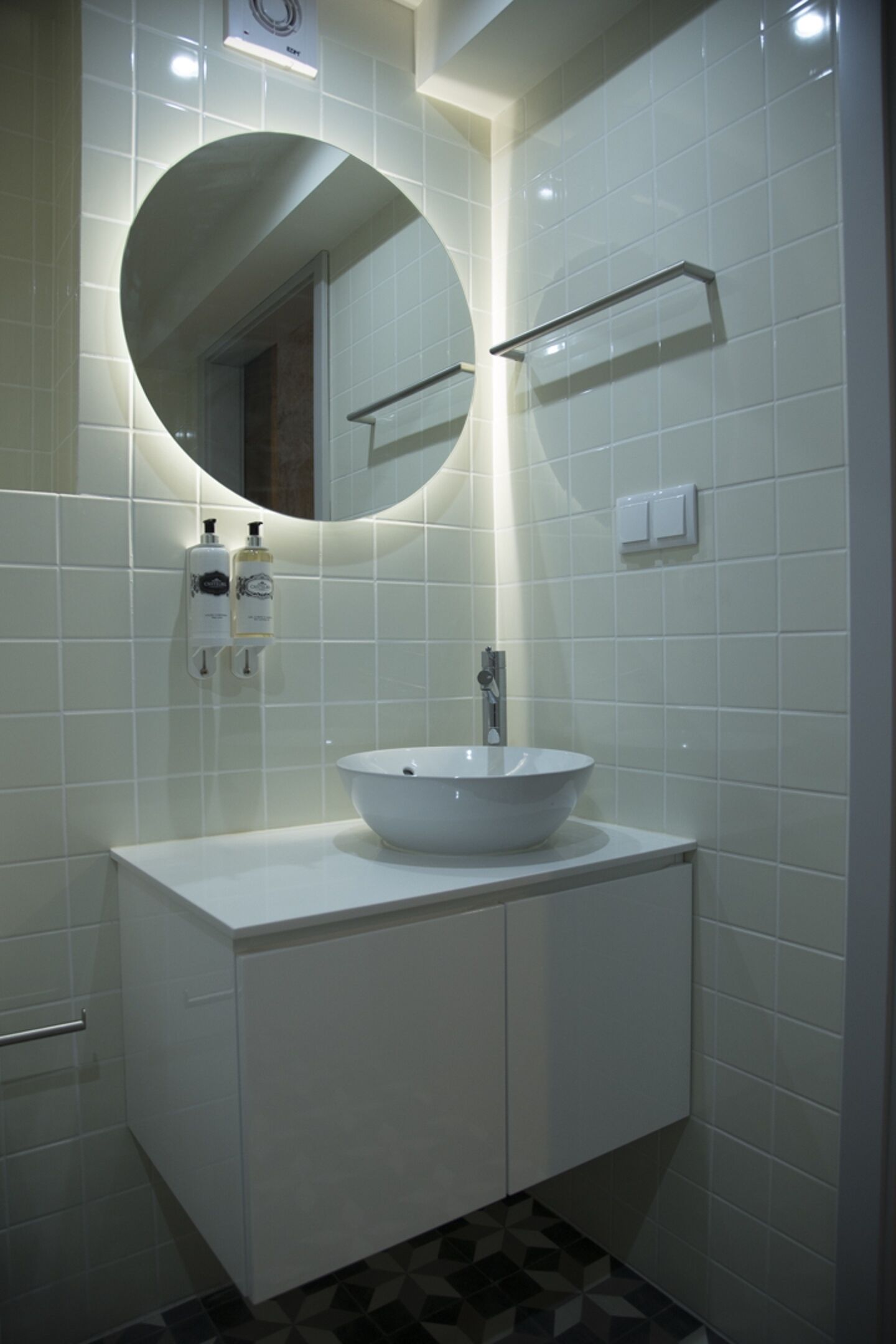 A modern bathroom with a round mirror, white tiles, a white sink, and a metal towel rack.