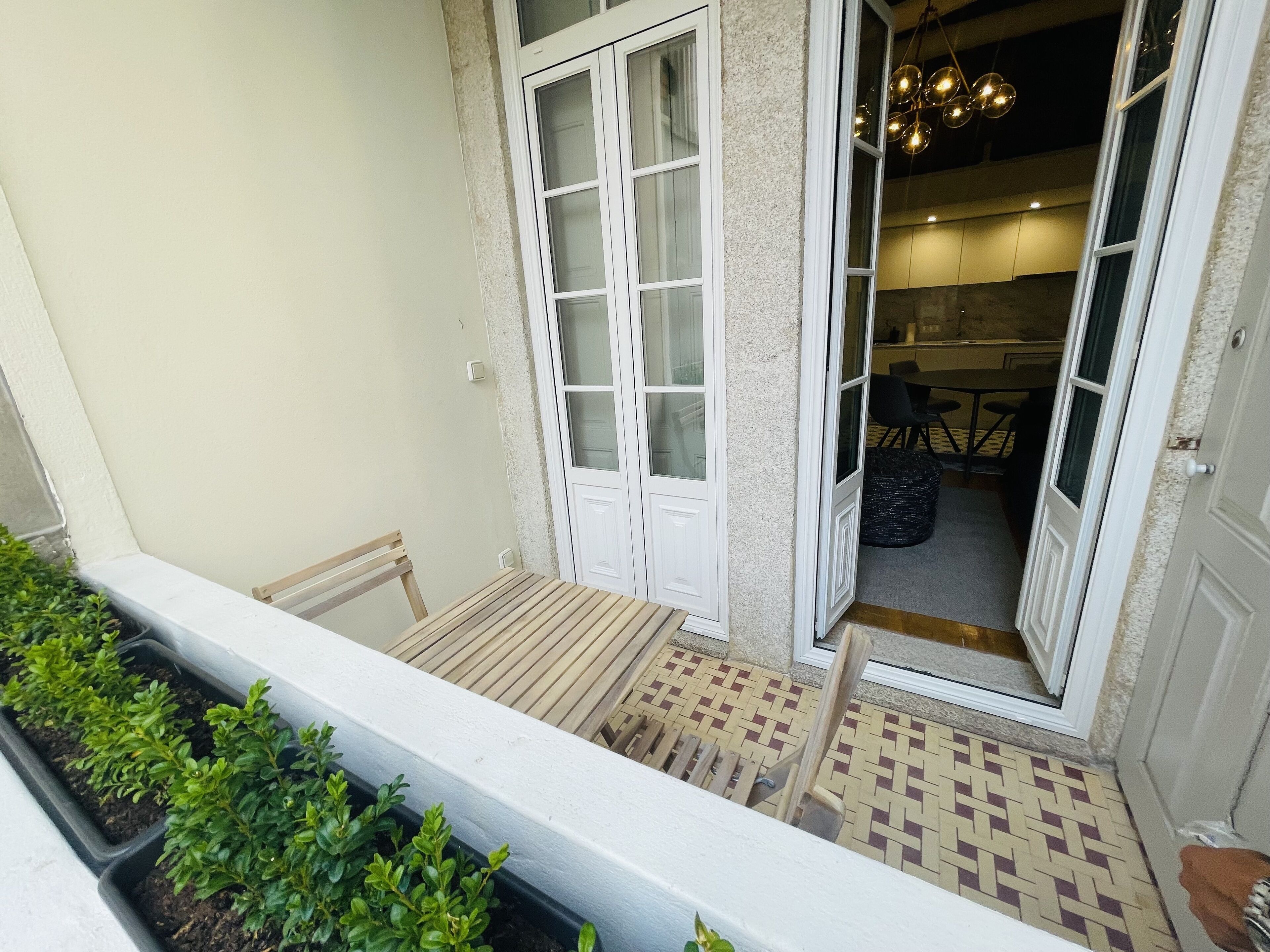 Un balcon avec une table et des chaises en bois, un petit jardin agrémenté de plantes en pot, et une vue sur l’intérieur d’une maison à travers des portes ouvertes.