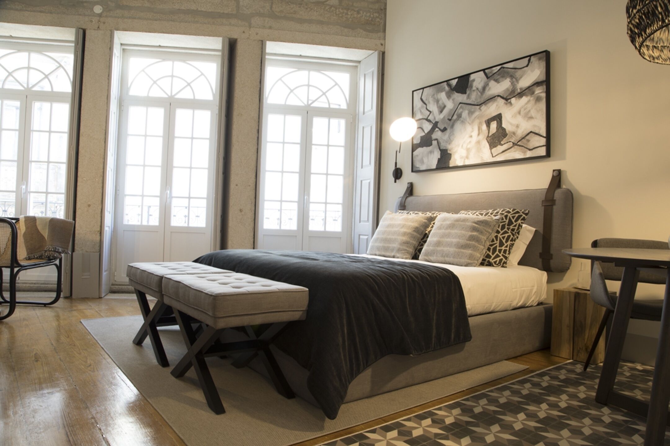 A bedroom with a large bed, a bench, a chair, and a table. There are windows with a view of the outside.
