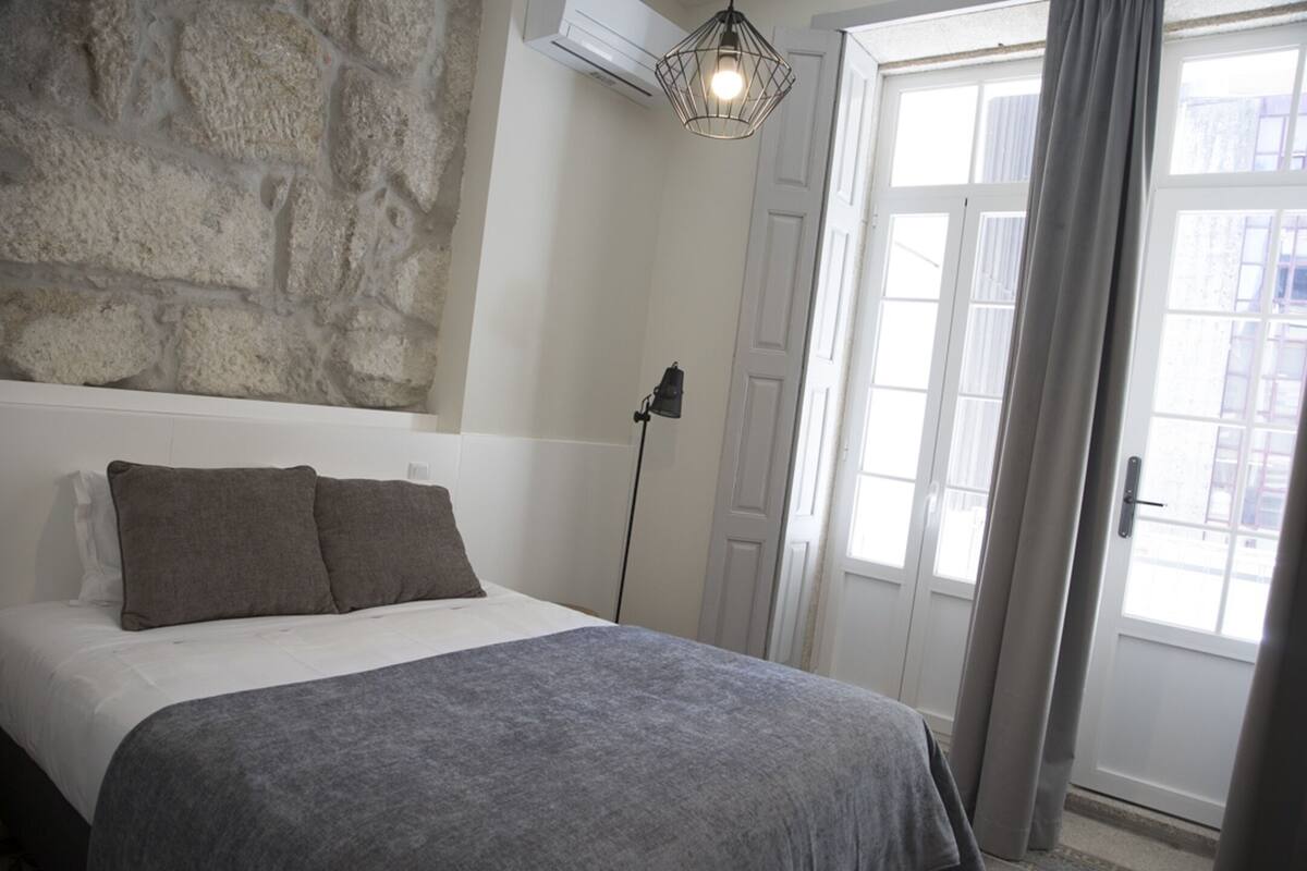 A bedroom with a stone wall, a bed with grey bedding, a floor lamp, and a door with curtains.