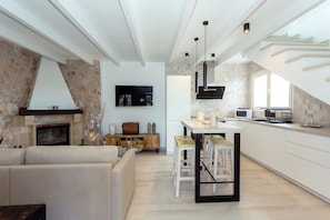 A modern kitchen with a stone wall, a fireplace, a dining area with stools, and a staircase leading to an upper level.