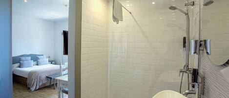 A modern bathroom with a glass-enclosed shower, a white sink, and a bed with a view of the room.