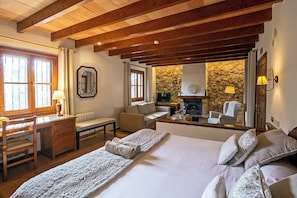 A bedroom with a large bed, wooden ceiling beams, a stone wall, a fireplace, and a seating area with a sofa and armchairs.