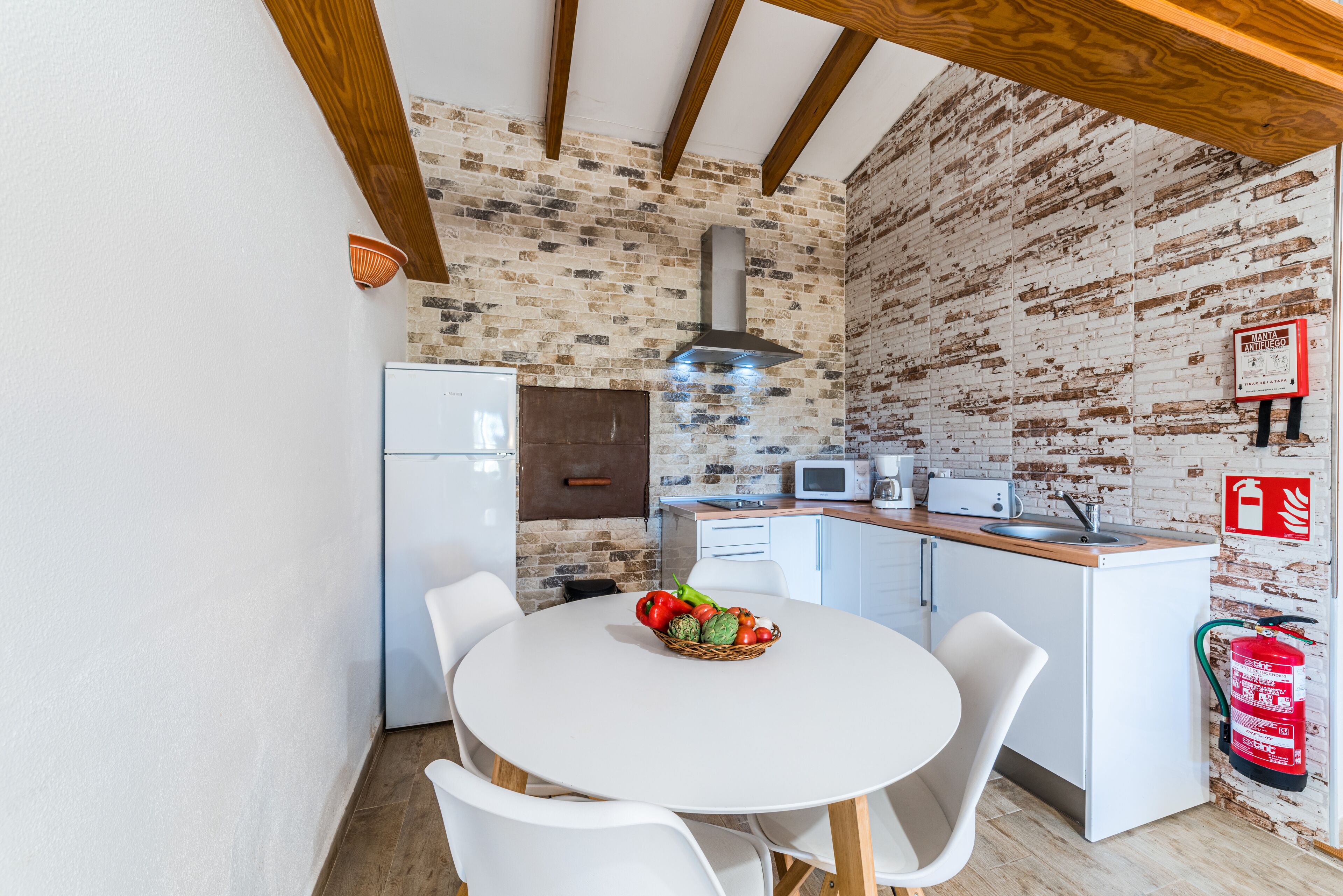 Une cuisine avec des meubles blancs, un mur en briques, une table blanche avec des chaises, un four à micro-ondes et un extincteur.