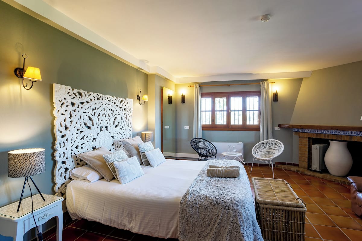 Une chambre à coucher avec un grand lit, une tête de lit décorative, des lampes fixées au mur, un fauteuil en osier et une cheminée.