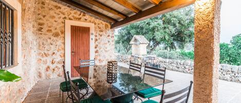 Un patio con paredes de piedra, una mesa de vidrio y sillas verdes acolchadas, una puerta de madera y un muro de piedra con un edificio pequeño al fondo.