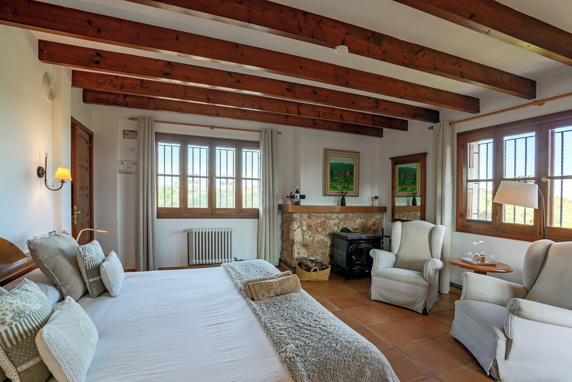 Een slaapkamer met een groot bed, twee fauteuils, een open haard en uitzicht op de omgeving.