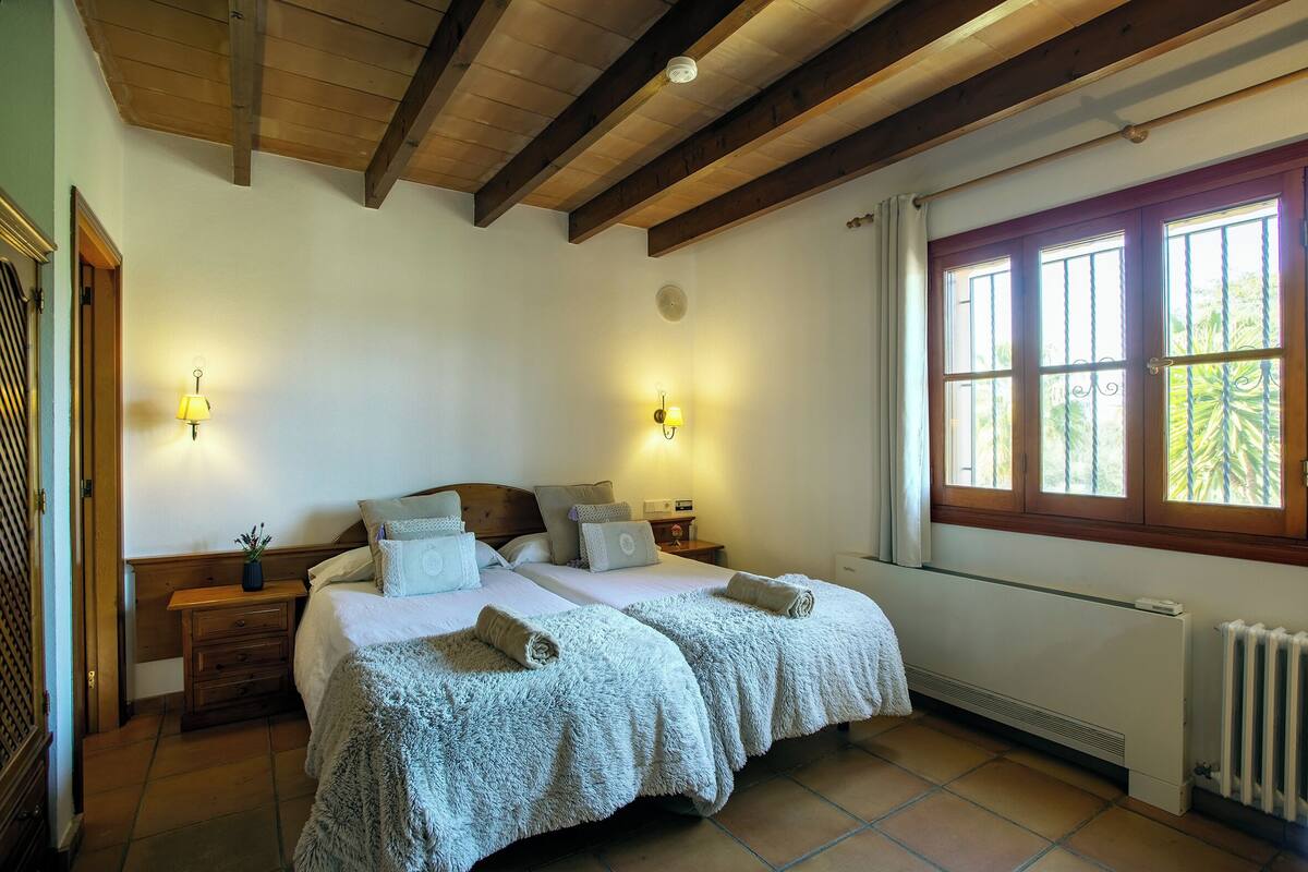 Une chambre avec deux lits, des poutres apparentes, deux lampes fixées au mur, une fenêtre avec vue et un radiateur.